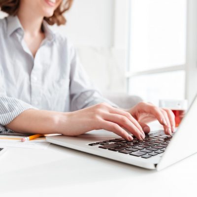 Cropped photo of pretty brunette woman typing email on laptop computer while sitting at home, selective focus on hand